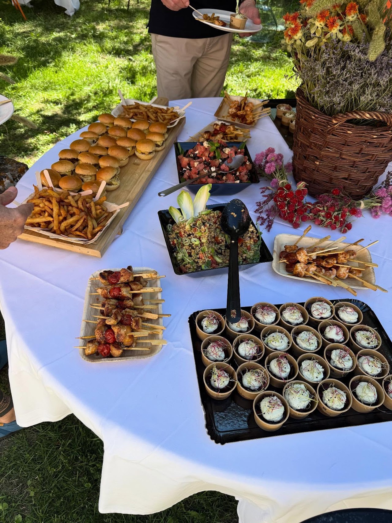 Buffet traiteur en plein air sur nappe blanche : mini-burgers, brochettes, verrines et salades.