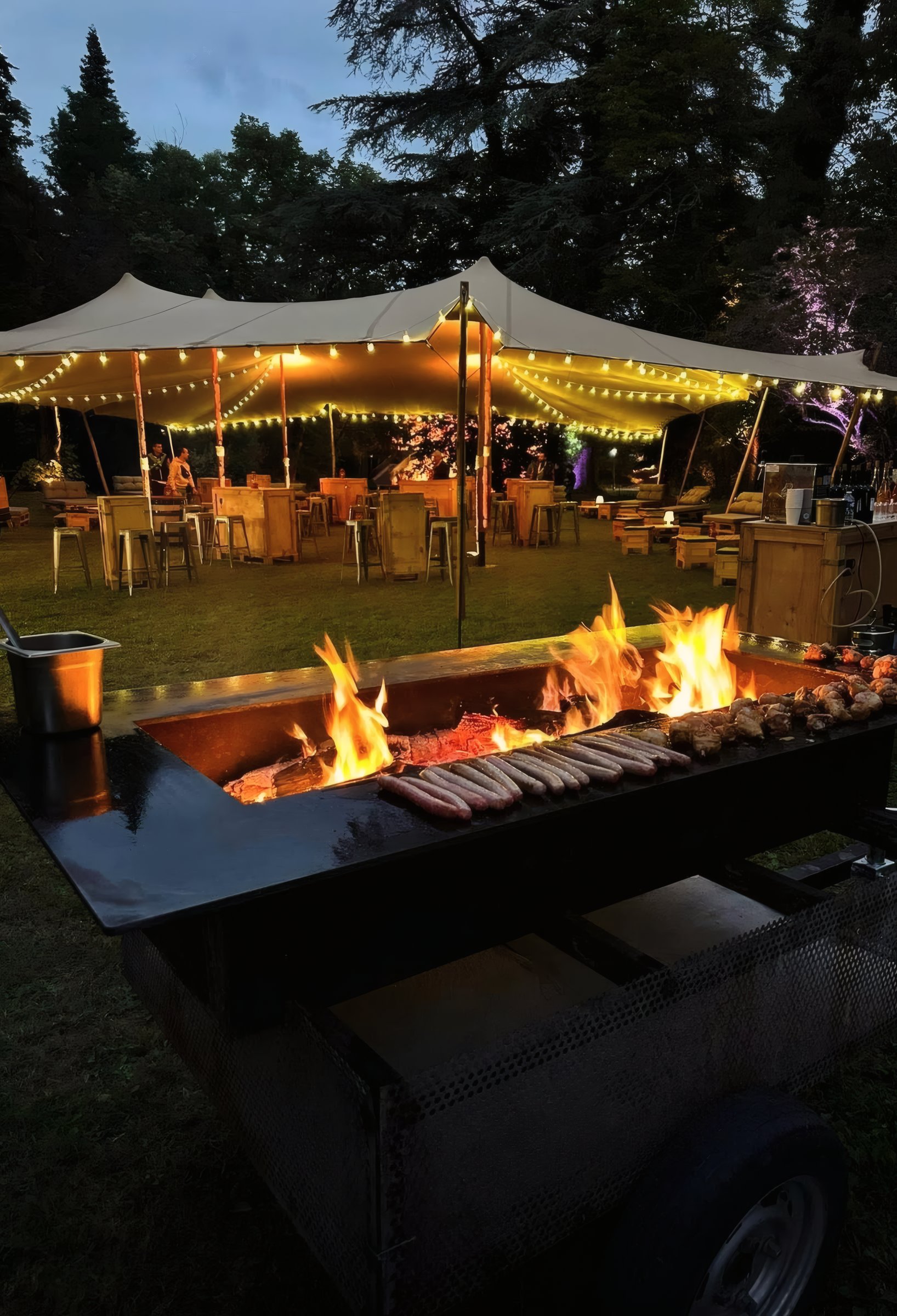 Plancha-brasero en flammes au crépuscule, grillades en cours sous une tente illuminée.
