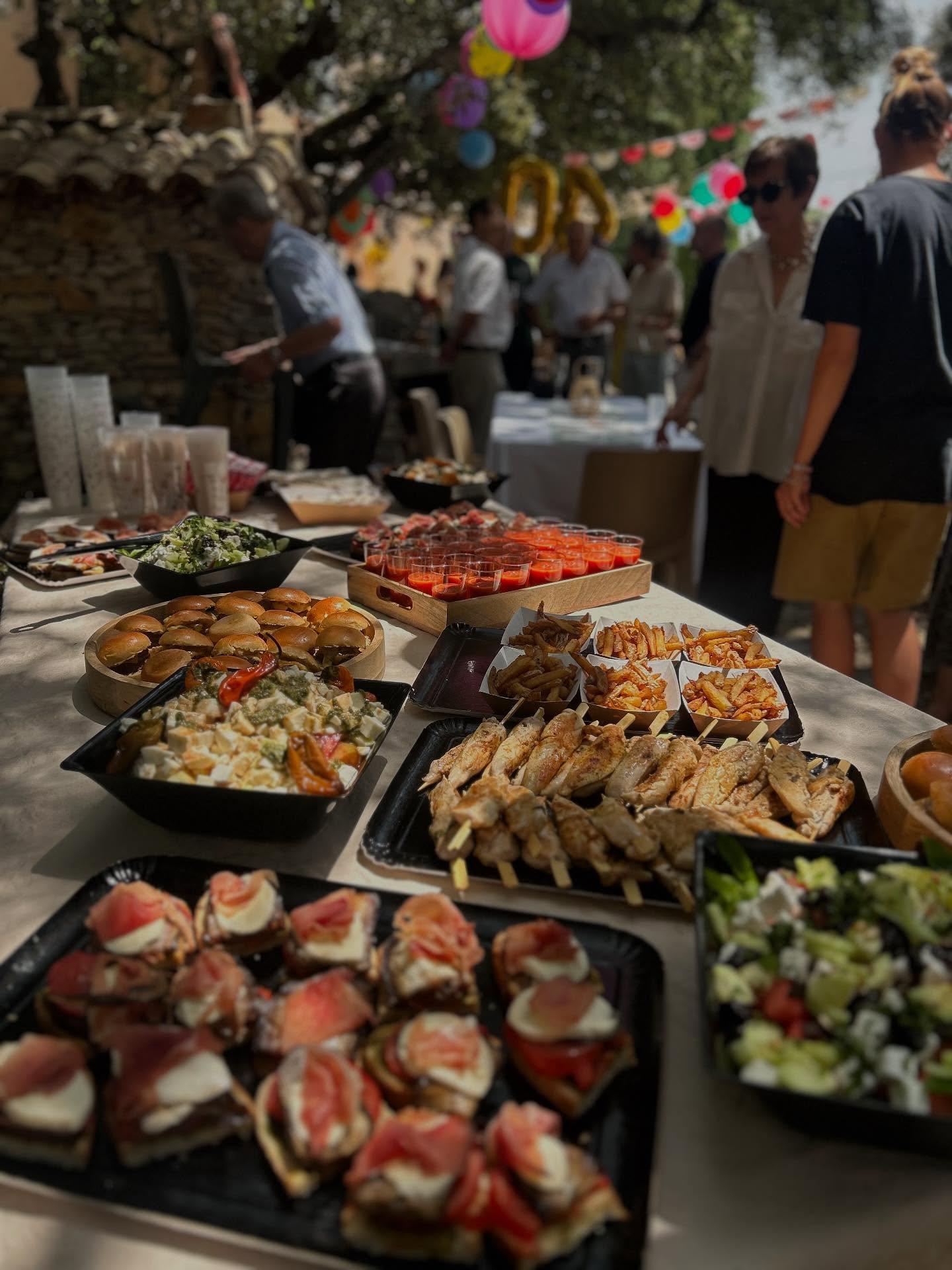 Buffet de réception en plein air sous les oliviers : mini-burgers, brochettes, salades, ambiance conviviale d'un événement privé.