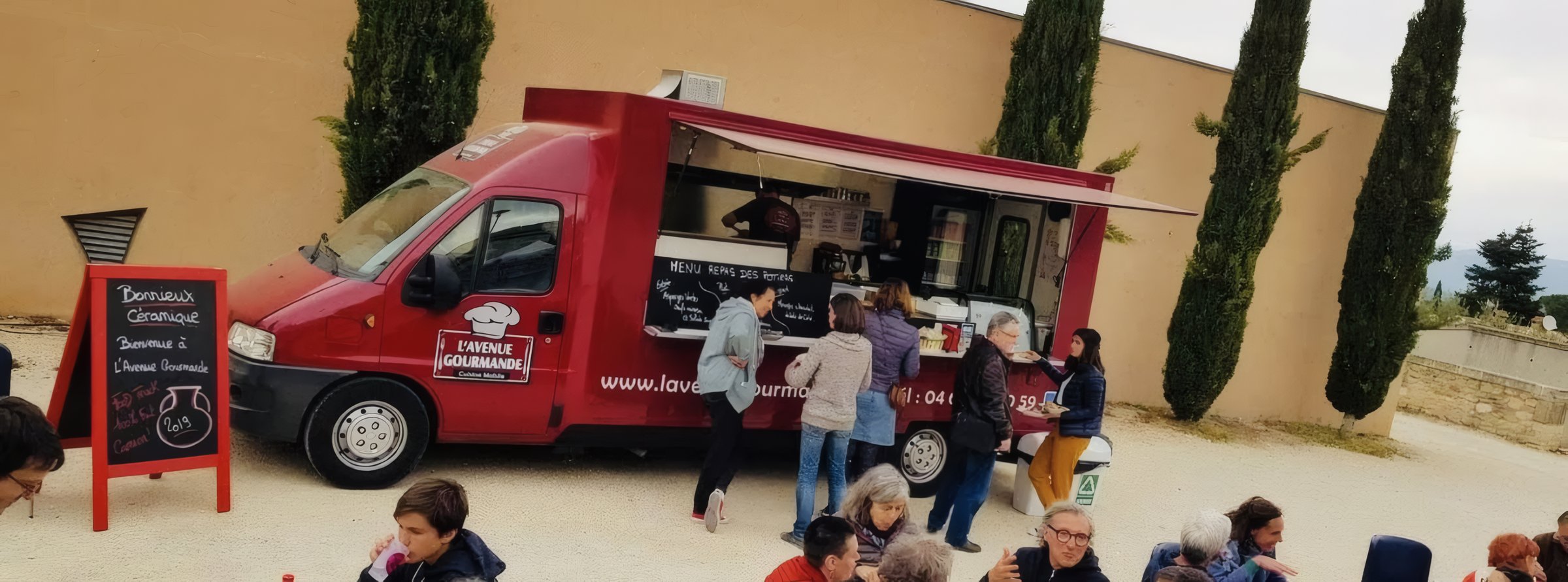 Le camion rouge de L'avenue gourmande, grand ouvert et en service, ardoise de menu et clients qui commandent devant un mas du Luberon.