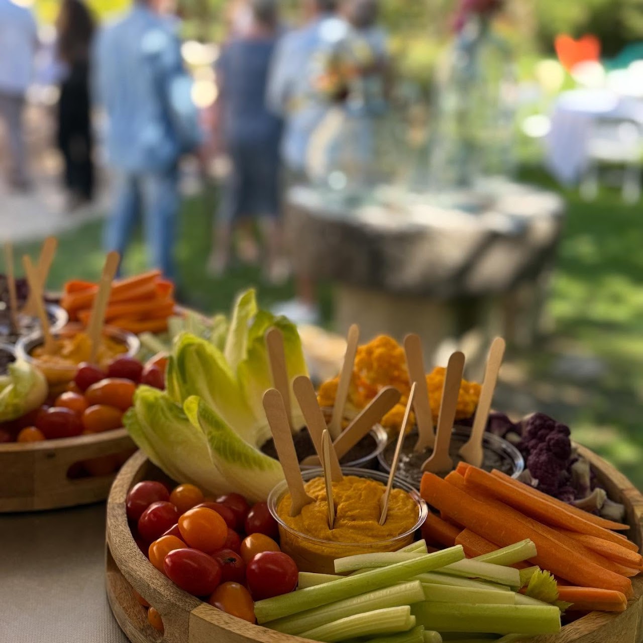 Plateaux de crudités et dips en extérieur, lumière estivale, convives en arrière-plan.