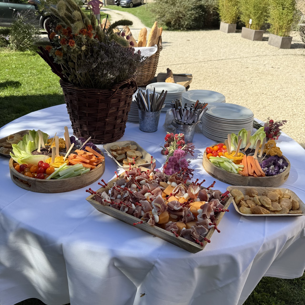Table de réception dressée en extérieur, nappe blanche, planches de charcuterie au melon et bouquet de fleurs séchées.